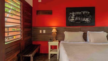 A hotel room with a bed, bedside table, lamp, and a window with a view of greenery.