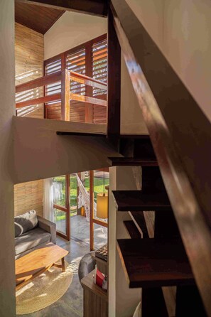 Interior de varios niveles con escalera, vigas de madera y vistas a un área exterior con sofá y mesa.