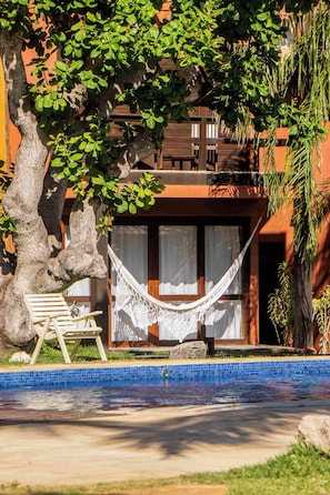 A poolside area with a hammock, a bench, and a view of a building with large windows. - Pousada Naquela (Jijoca de Jericoacoara)