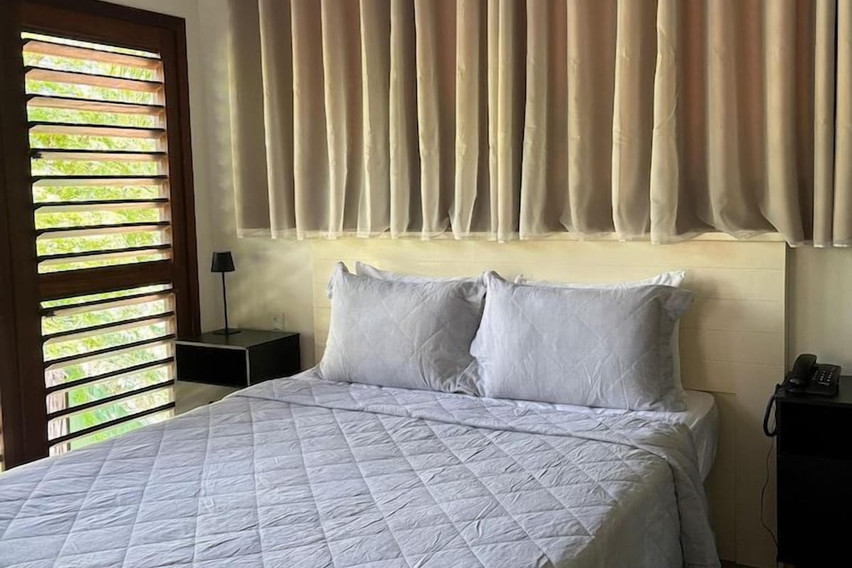 Un dormitorio con una cama grande, cortinas color beige y una ventana con persianas.