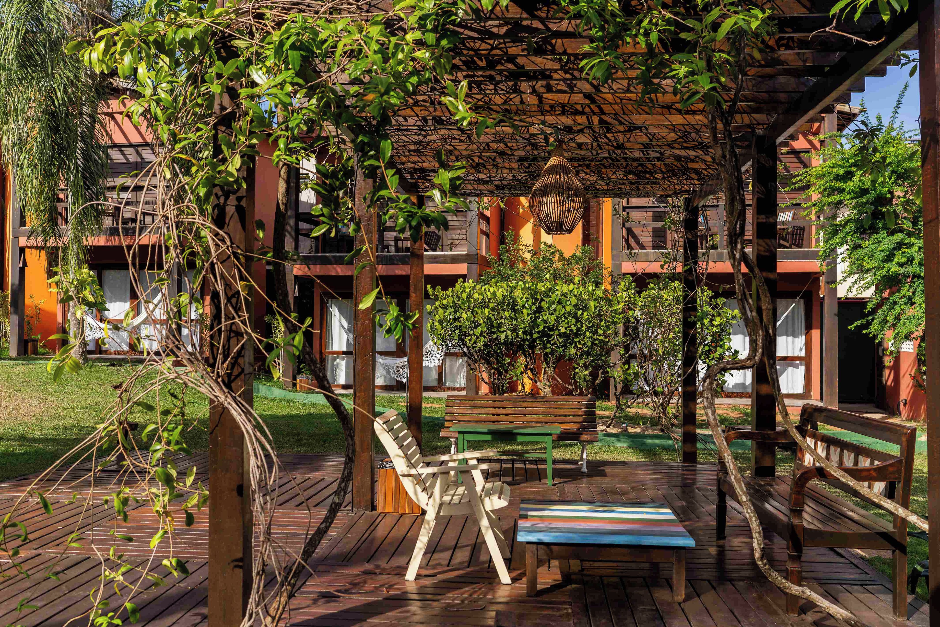 Una terraza de madera con un banco y una mesa, rodeada de follaje y una pérgola de madera.