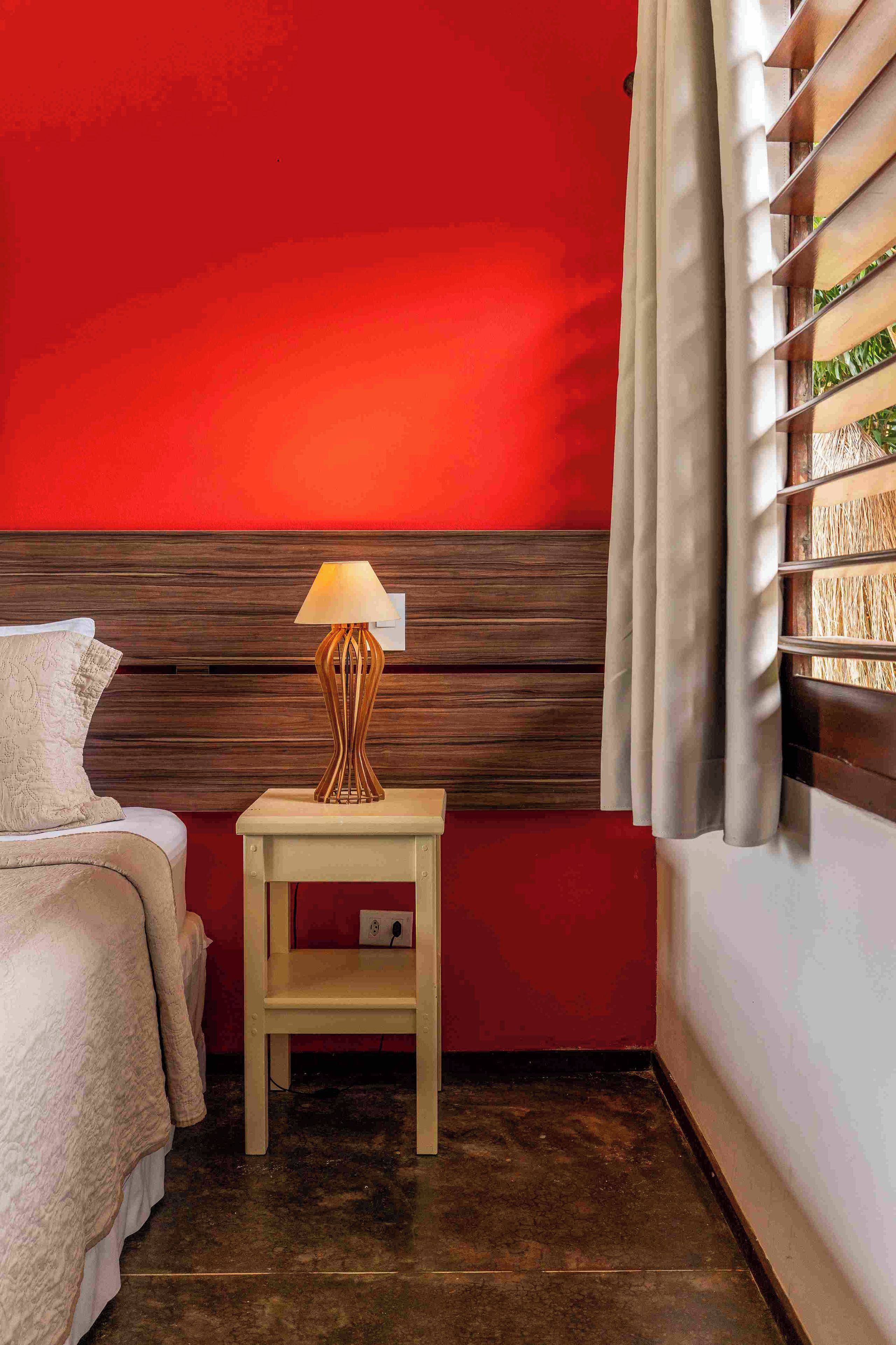 A hotel room with a red wall, a wooden headboard, a bedside table with a lamp, a bed with a white cover, and a window with blinds.