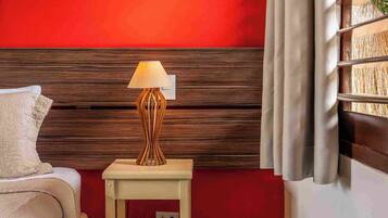 A hotel room with a red wall, a wooden headboard, a bedside table with a lamp, a bed with a white cover, and a window with blinds.