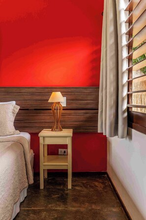 A hotel room with a red wall, a wooden headboard, a bedside table with a lamp, a bed with a white cover, and a window with blinds. - Pousada Naquela (Jijoca de Jericoacoara)
