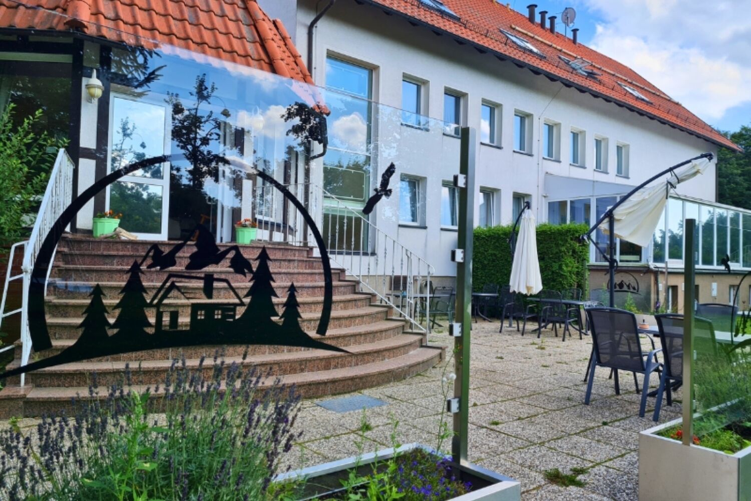 A two-story building with a red-tiled roof, outdoor seating area with tables and chairs, and a garden with plants and a decorative metal sculpture.