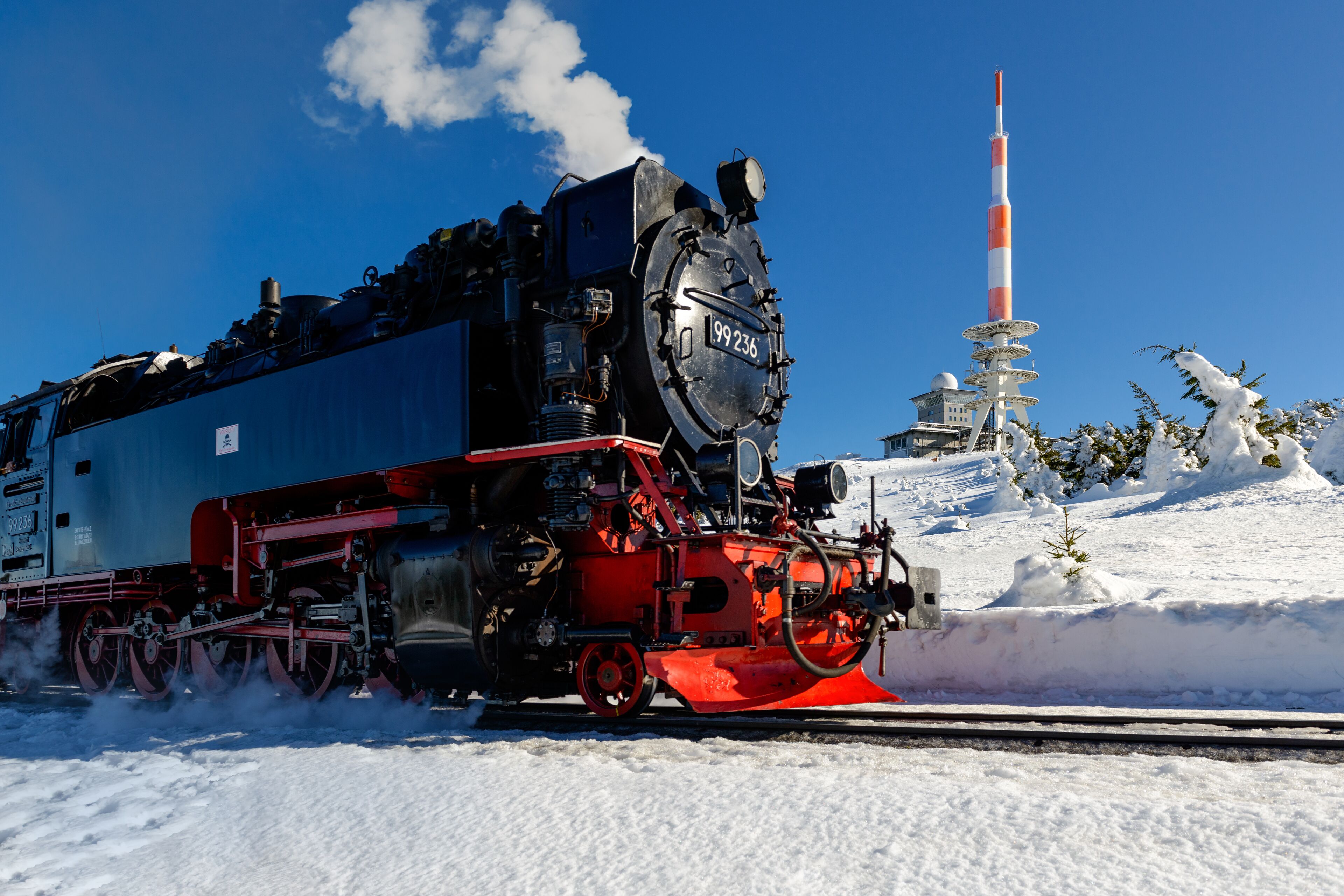 Eine Dampflokomotive mit auff älligem Nummernschild fährt auf Gleisen durch eine verschneite Landschaft, im Hintergrund ragt ein hoher, gestreifter Turm auf.