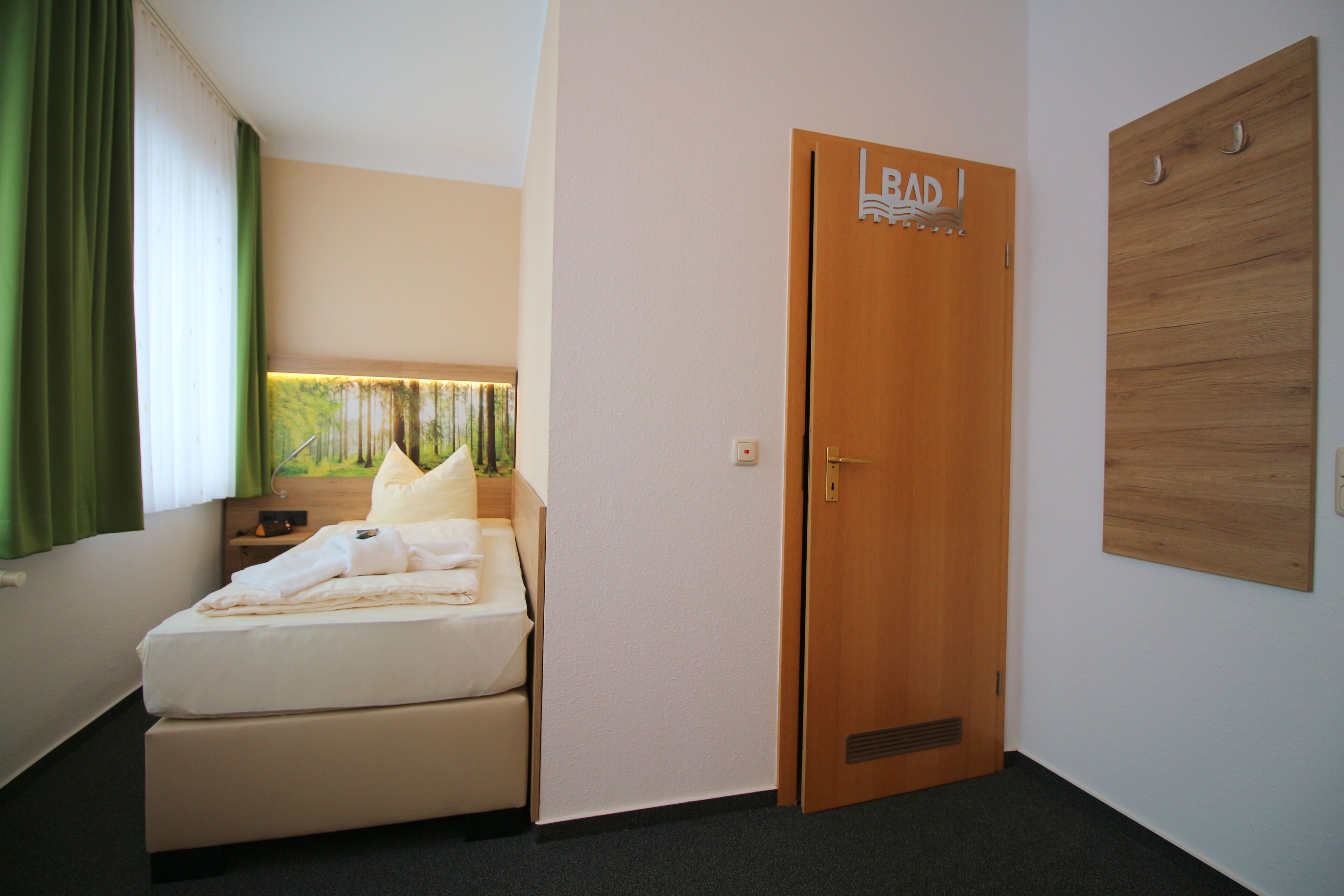 A hotel room with a bed, a nightstand, a green curtain, a painting, a wooden door with a sign, and a wooden panel on the wall.