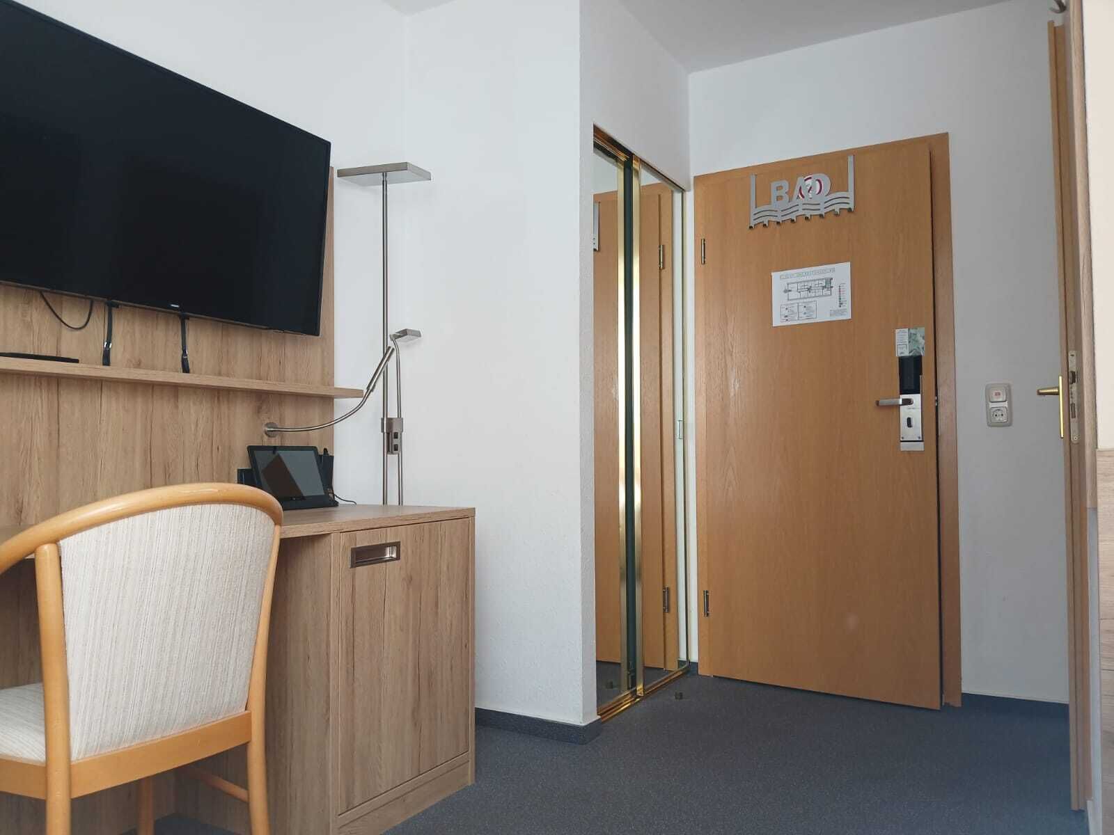 A hotel room with a wooden desk, a chair, a flat-screen TV, a floor lamp, and a door with a sign.