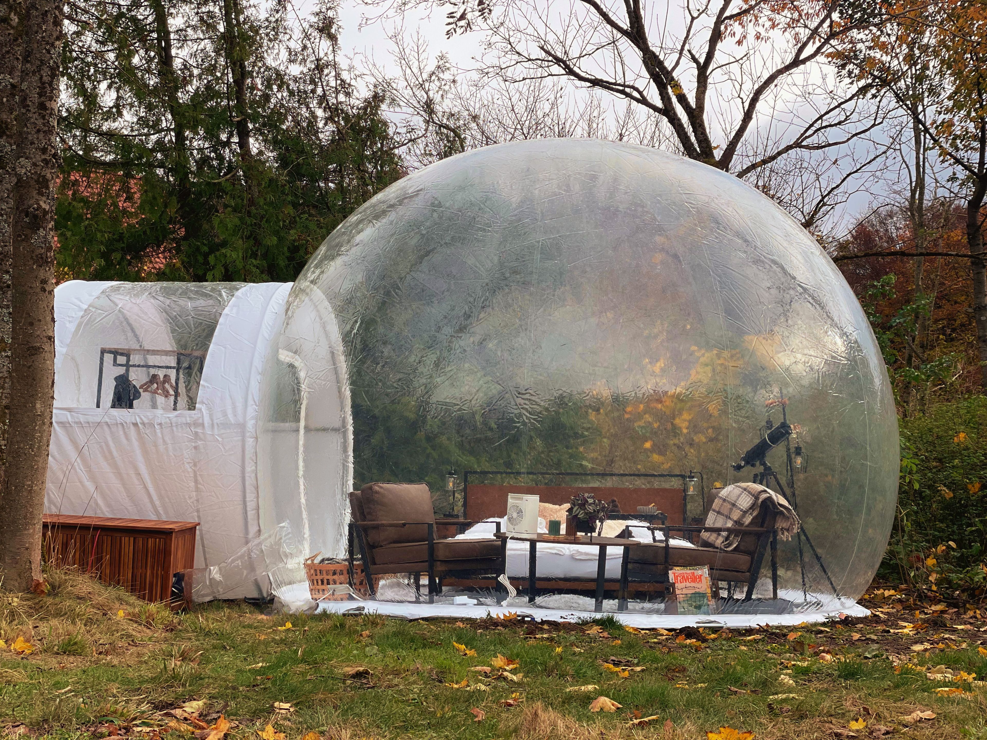 A transparent dome structure with a bed and a small table inside, surrounded by trees and grass.