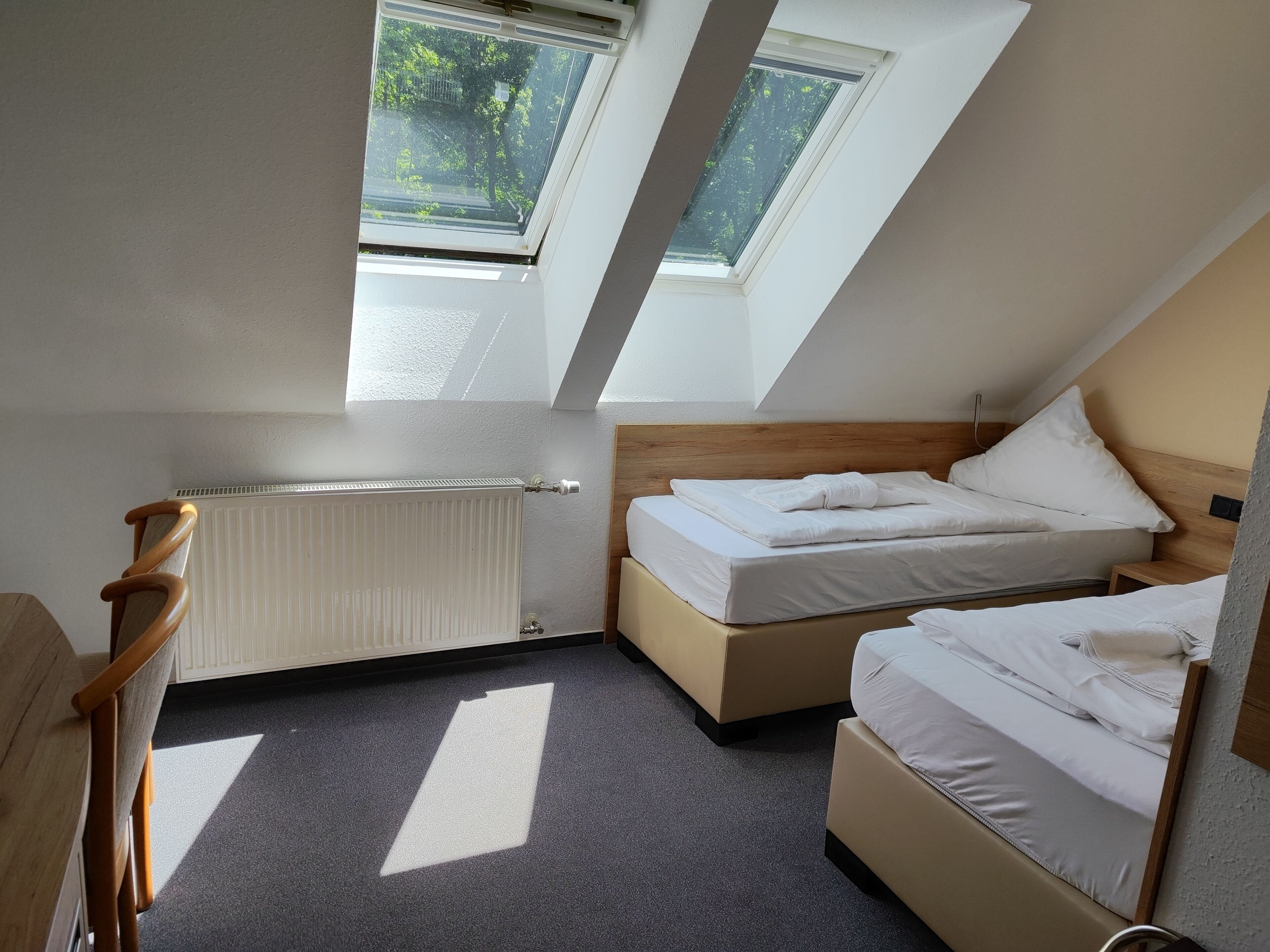 A small, well-lit room with a sloped ceiling, two beds, a wooden chair, a radiator, and skylights.