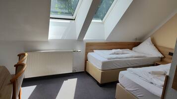A small, well-lit room with a sloped ceiling, two beds, a wooden chair, a radiator, and skylights.