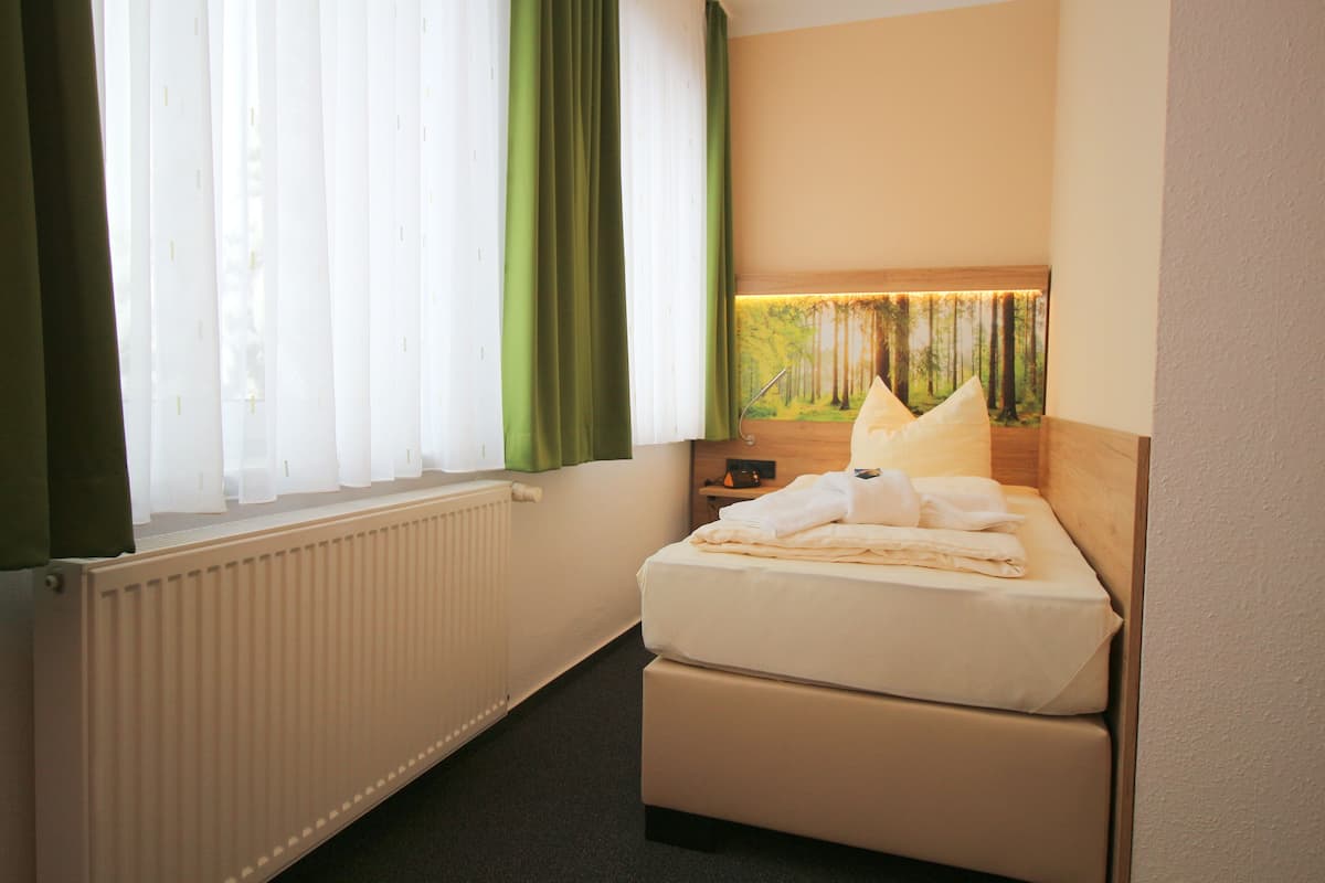 Habitación de hotel con cama, radiador, ventana con cortinas y una lámpara fijada a la pared.