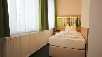 A hotel room with a bed, a radiator, a window with curtains, and a wall-mounted lamp.