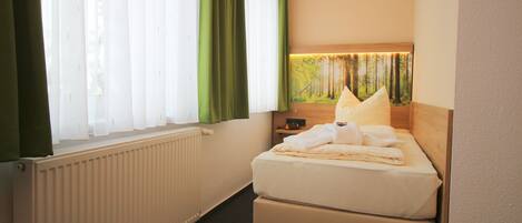 Habitación de hotel con cama, radiador, ventana con cortinas y una lámpara fijada a la pared.