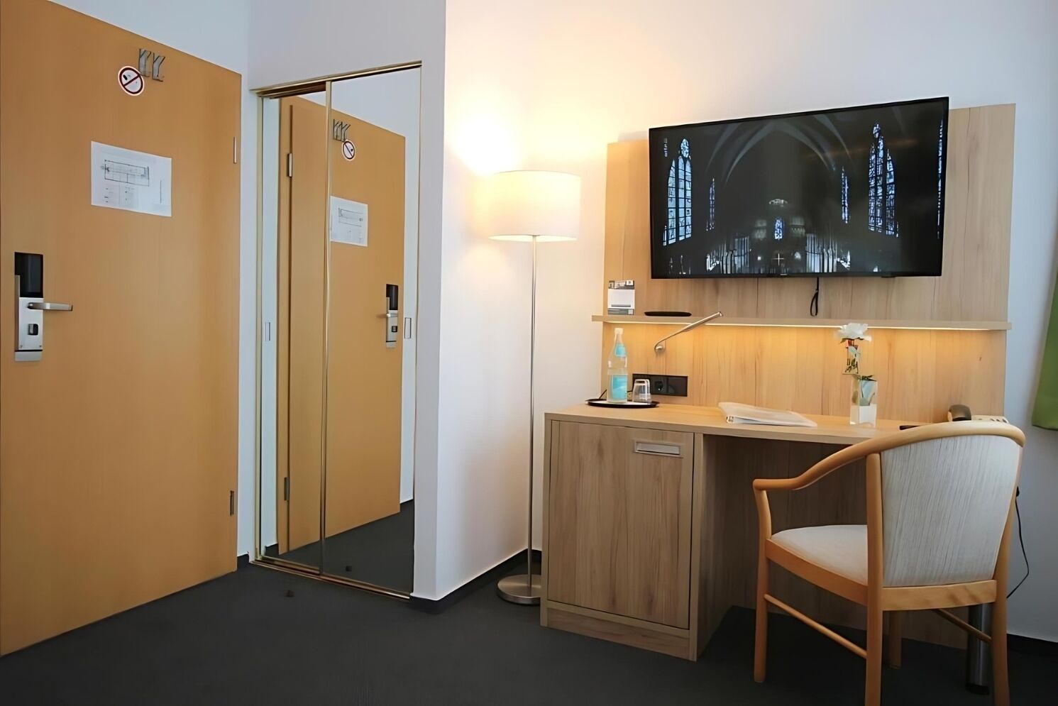 Une chambre d’hôtel avec un bureau en bois, une chaise, un téléviseur à écran plat fixé au mur et une porte dotée d’un miroir.