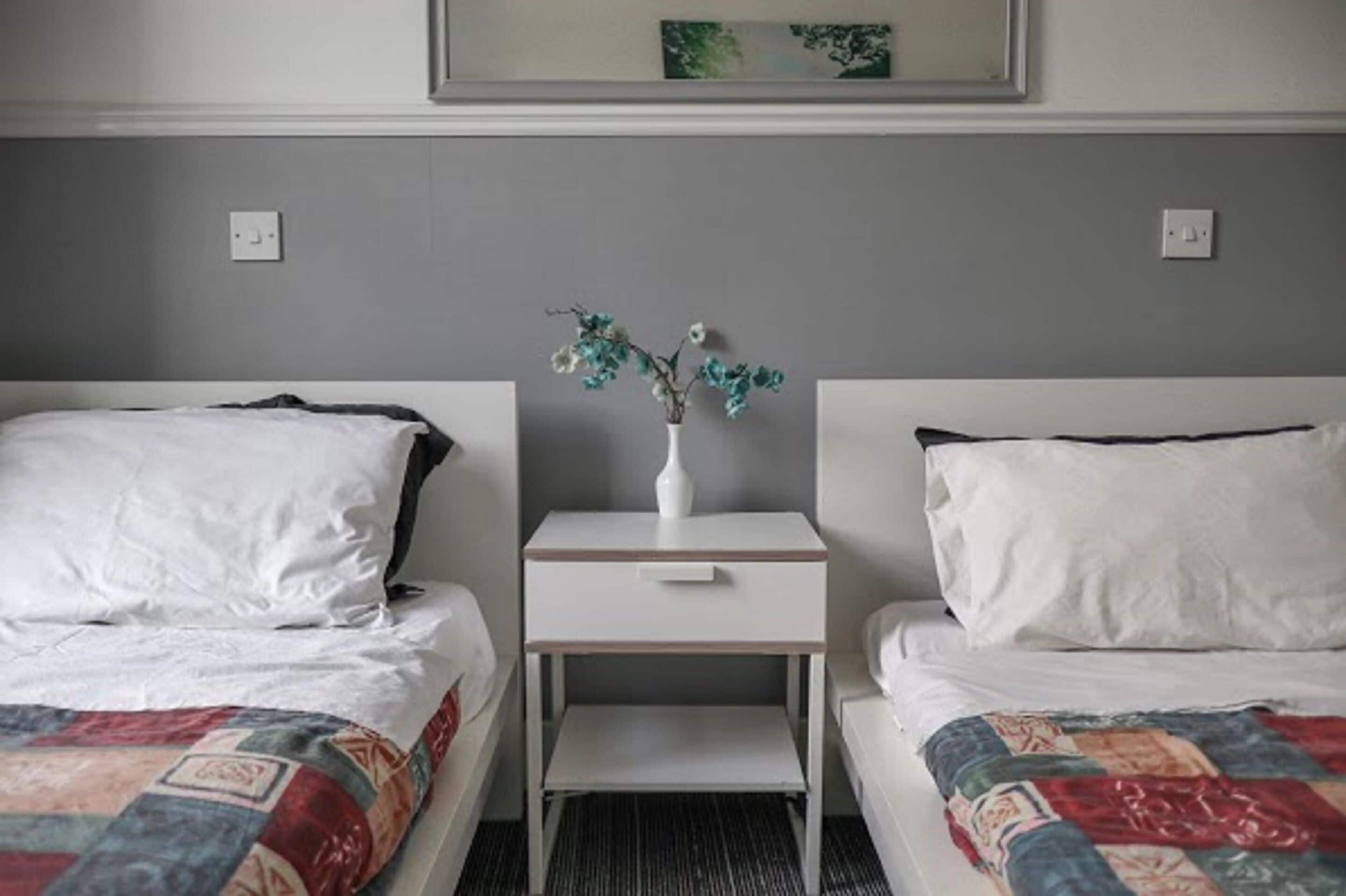 A hotel room with two single beds, a small bedside table with a vase of flowers, and a framed picture on the wall.