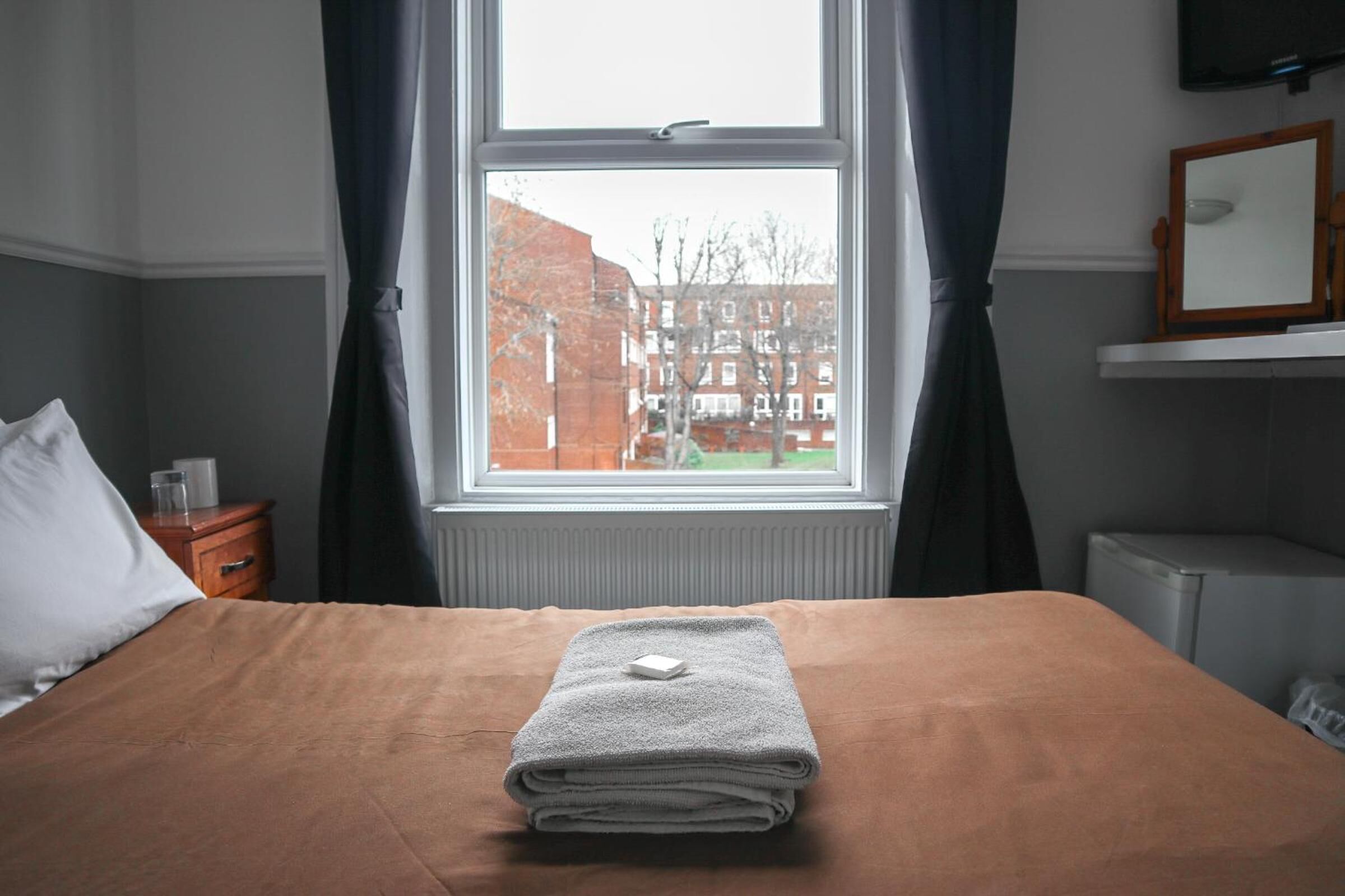 Una cama bien hecha con una toalla doblada y un objeto pequeño encima, una ventana con cortinas, una mesita de noche de madera con un vaso y un espejo en la pared.