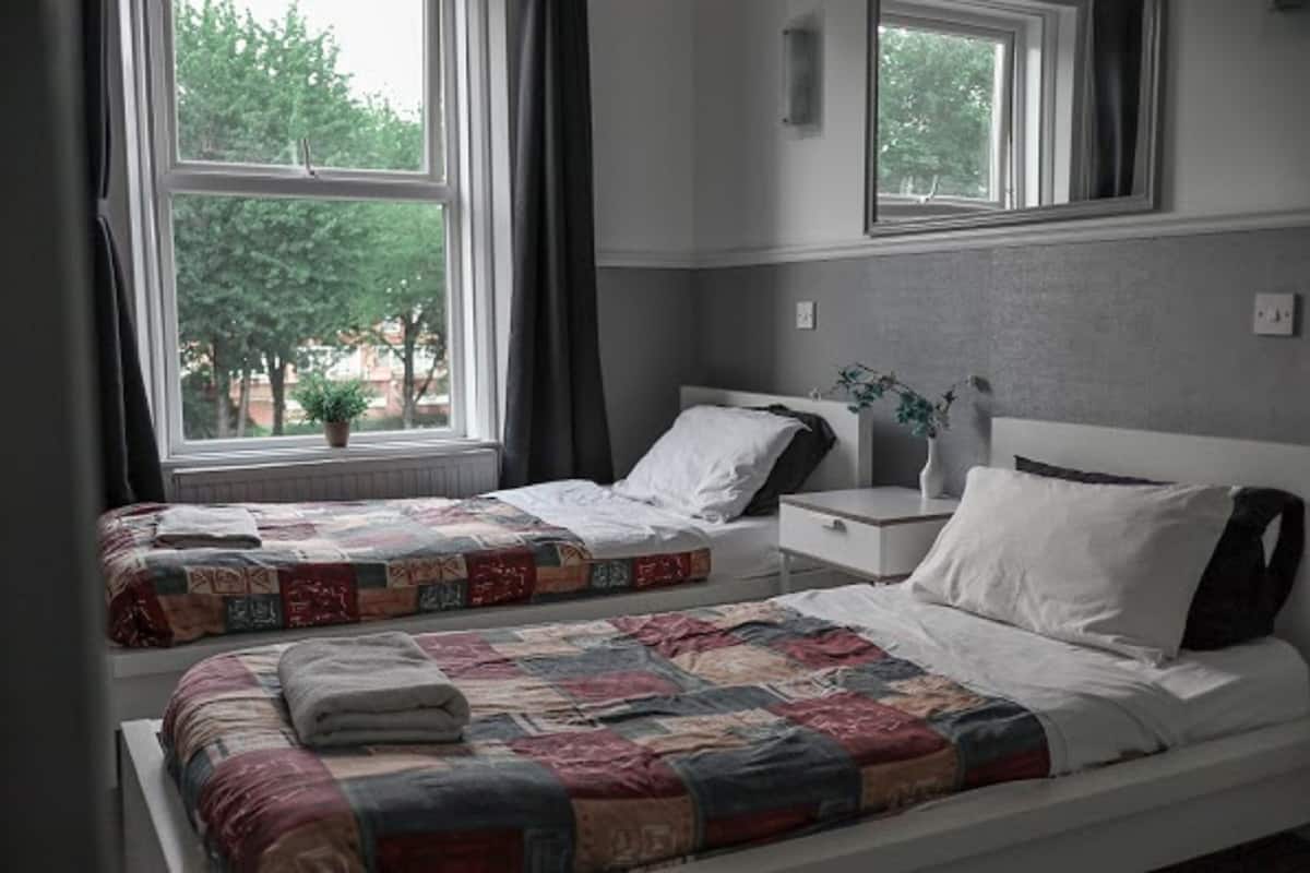 A bedroom with two beds, a nightstand, a window with curtains, and a view of trees outside.