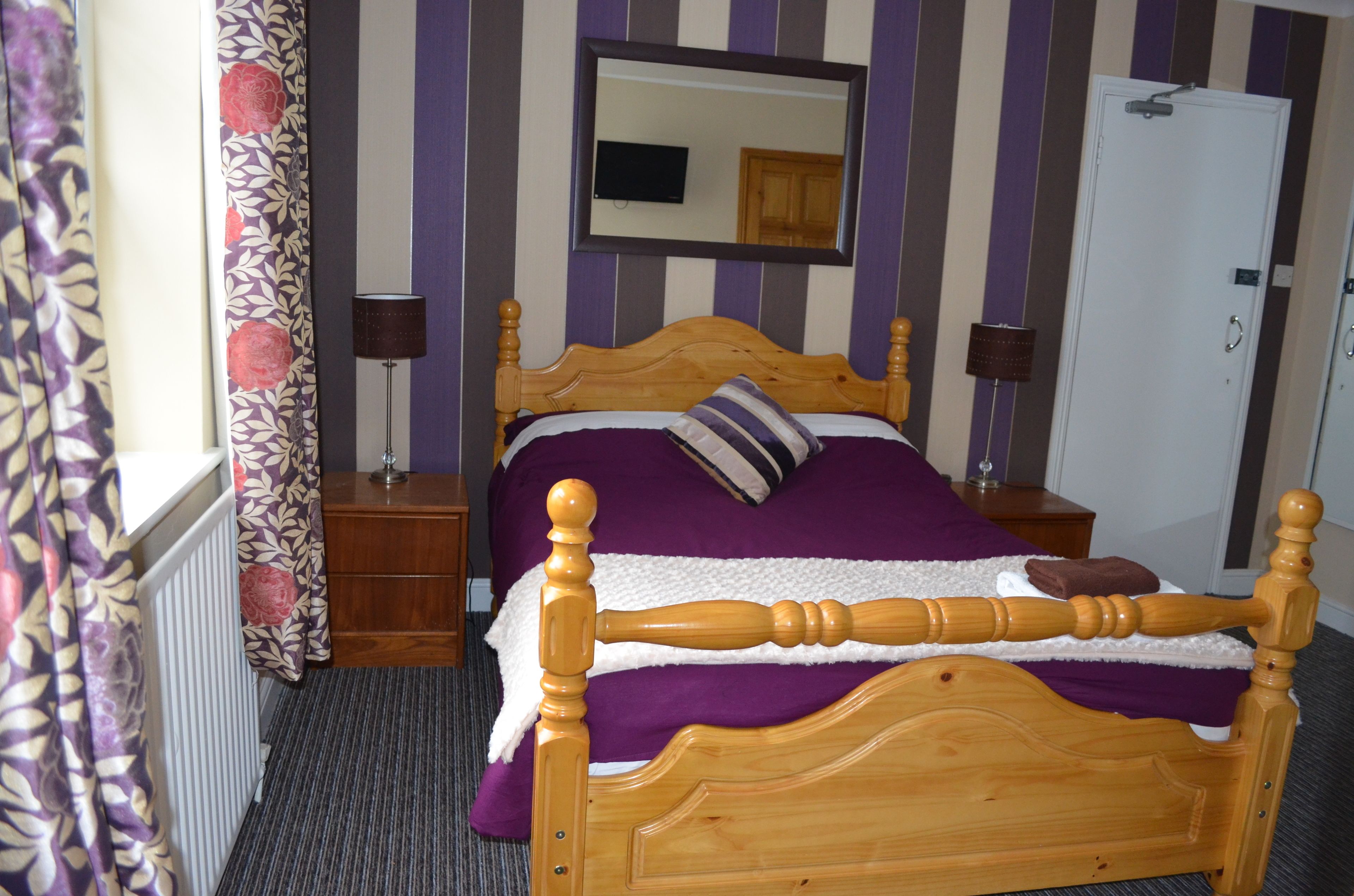 A wooden bed with a headboard, a bedside table, a lamp, a mirror, and a door in a room with striped walls.