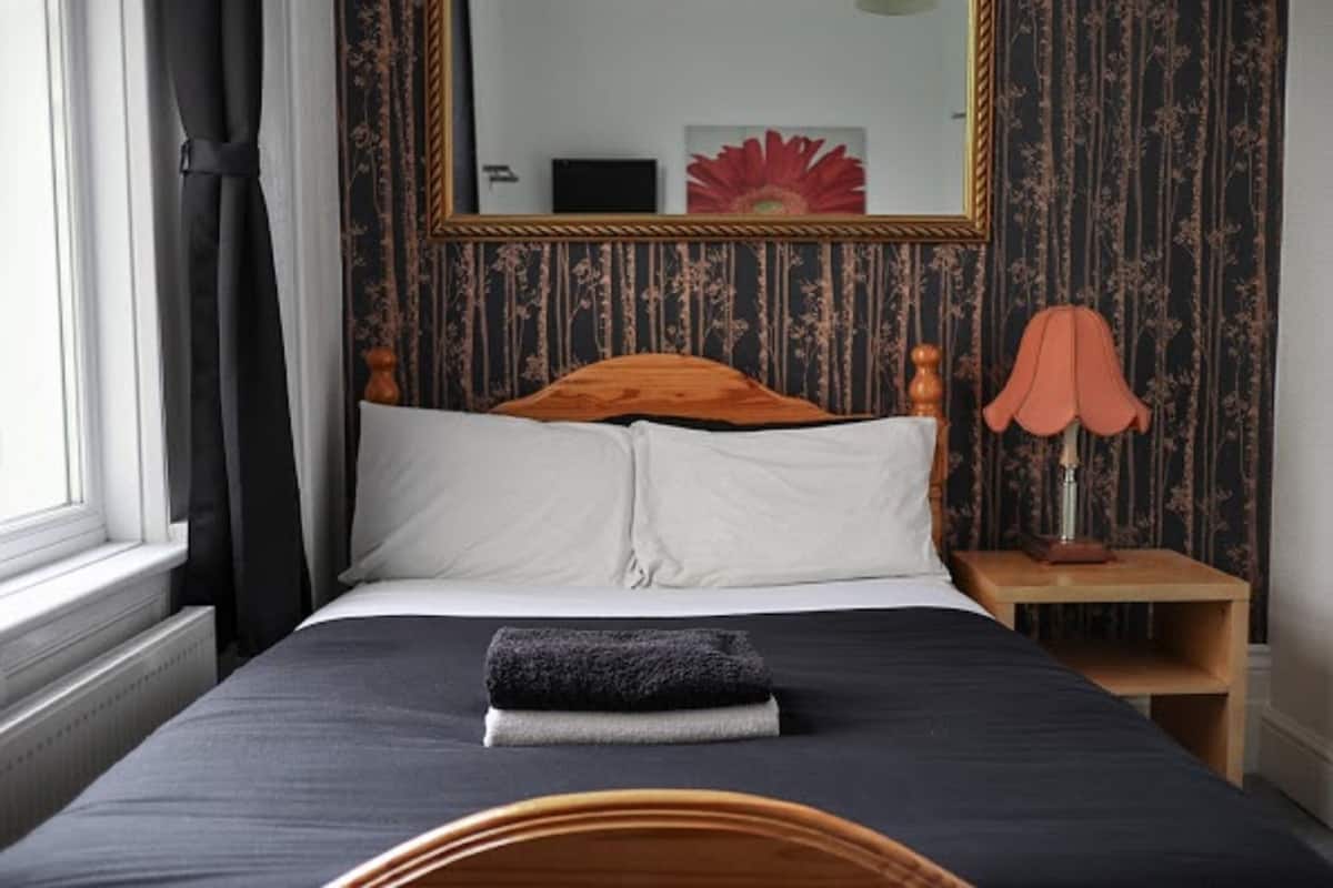 A neatly made bed with a dark bedspread and white pillows, a wooden headboard, a nightstand with a lamp, and a large mirror on the wall.