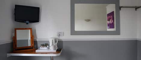 A bathroom with a mounted television, a mirror, a small table with a kettle and cup, and a towel rack.