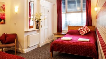 Hotellihuone, jossa on sänky, tuoli, pieni pöytä, ikkuna verhoilla ja patteri.