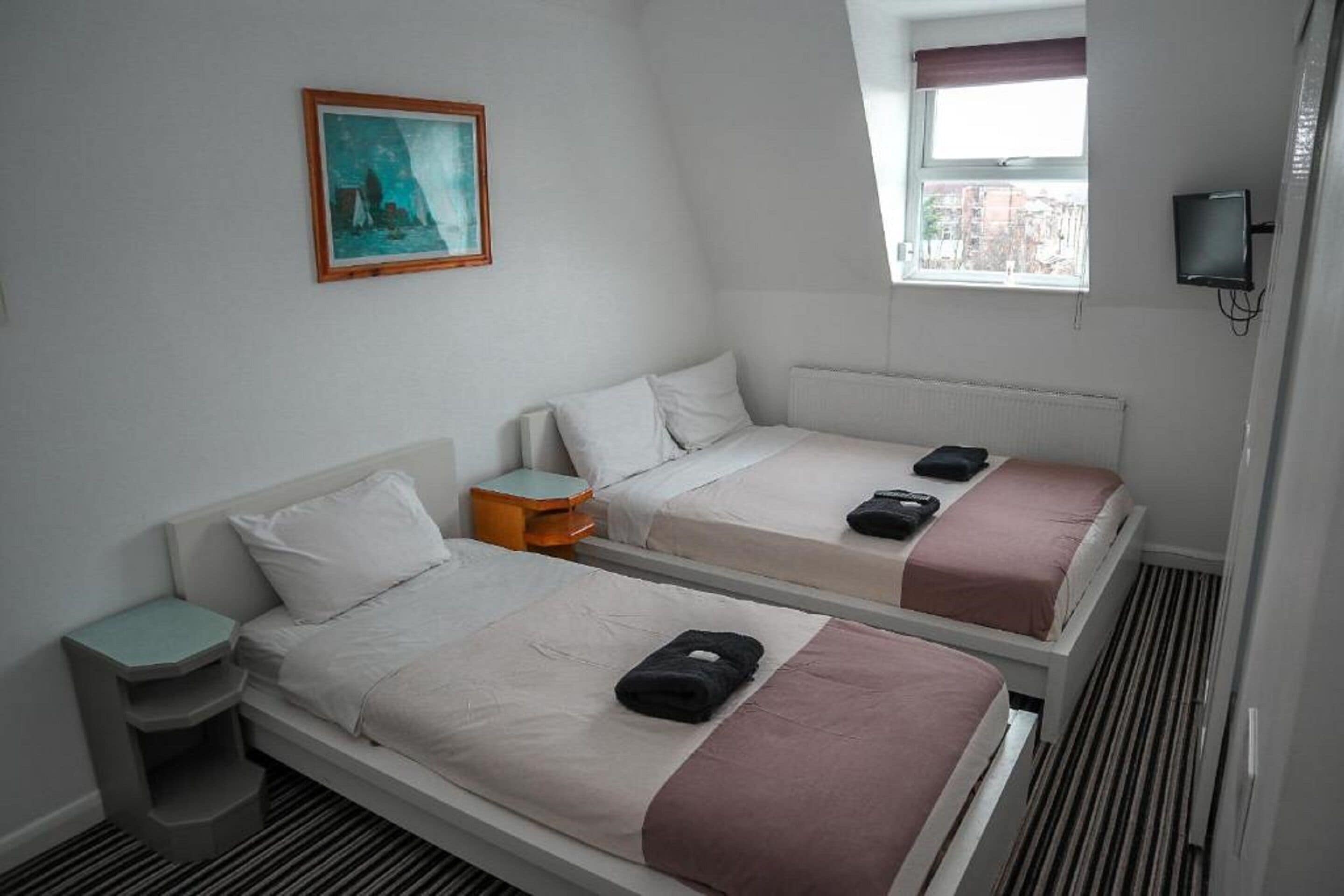 A hotel room with two single beds, a small table, a nightstand, a painting on the wall, a window with a view, and a television.