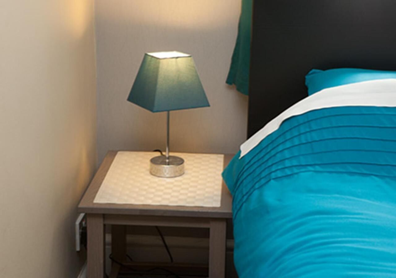 A neatly made bed with a teal comforter and a white pillow, a small bedside table with a lamp, and a green towel hanging on the wall.