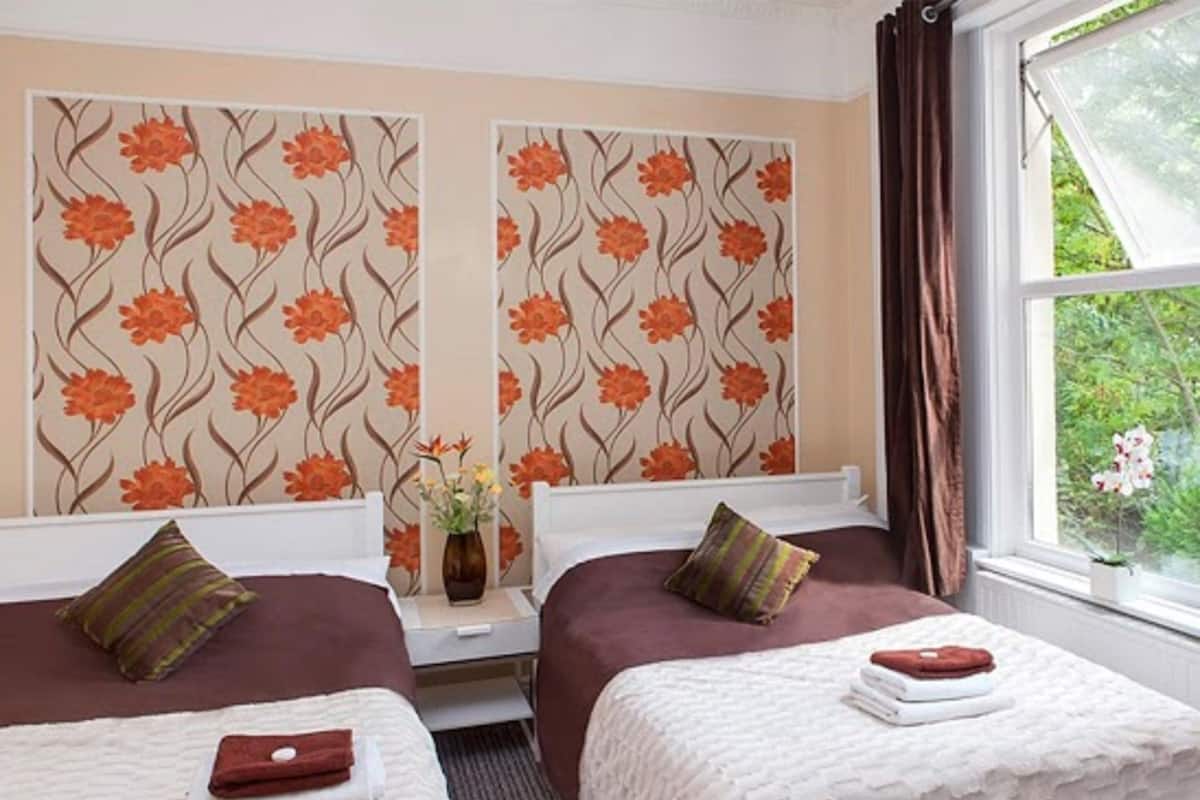 A hotel room with two beds, a window with curtains, and a floral wallpaper design.