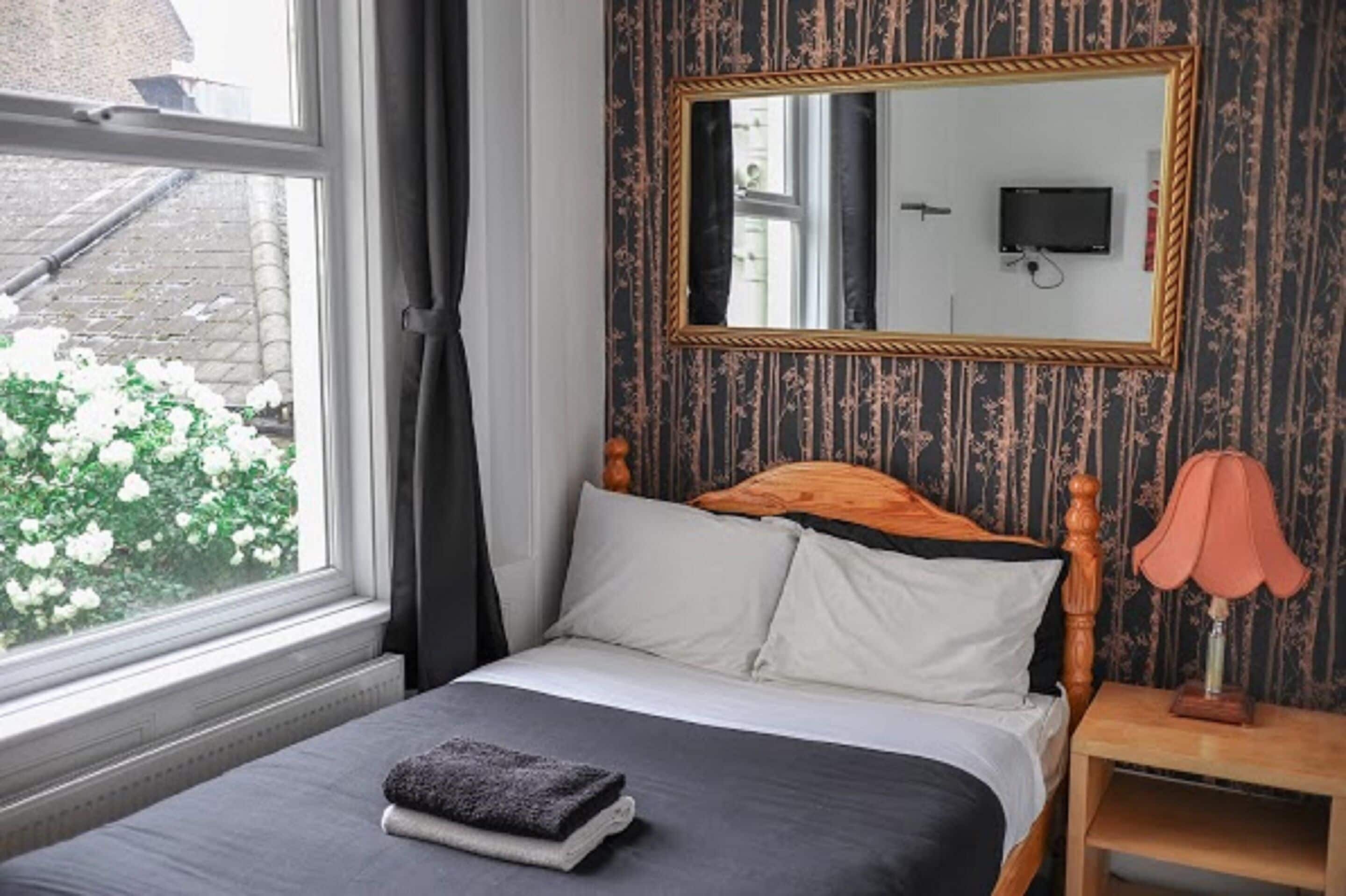 A bedroom with a bed, a nightstand, a lamp, a mirror, and a window with curtains.