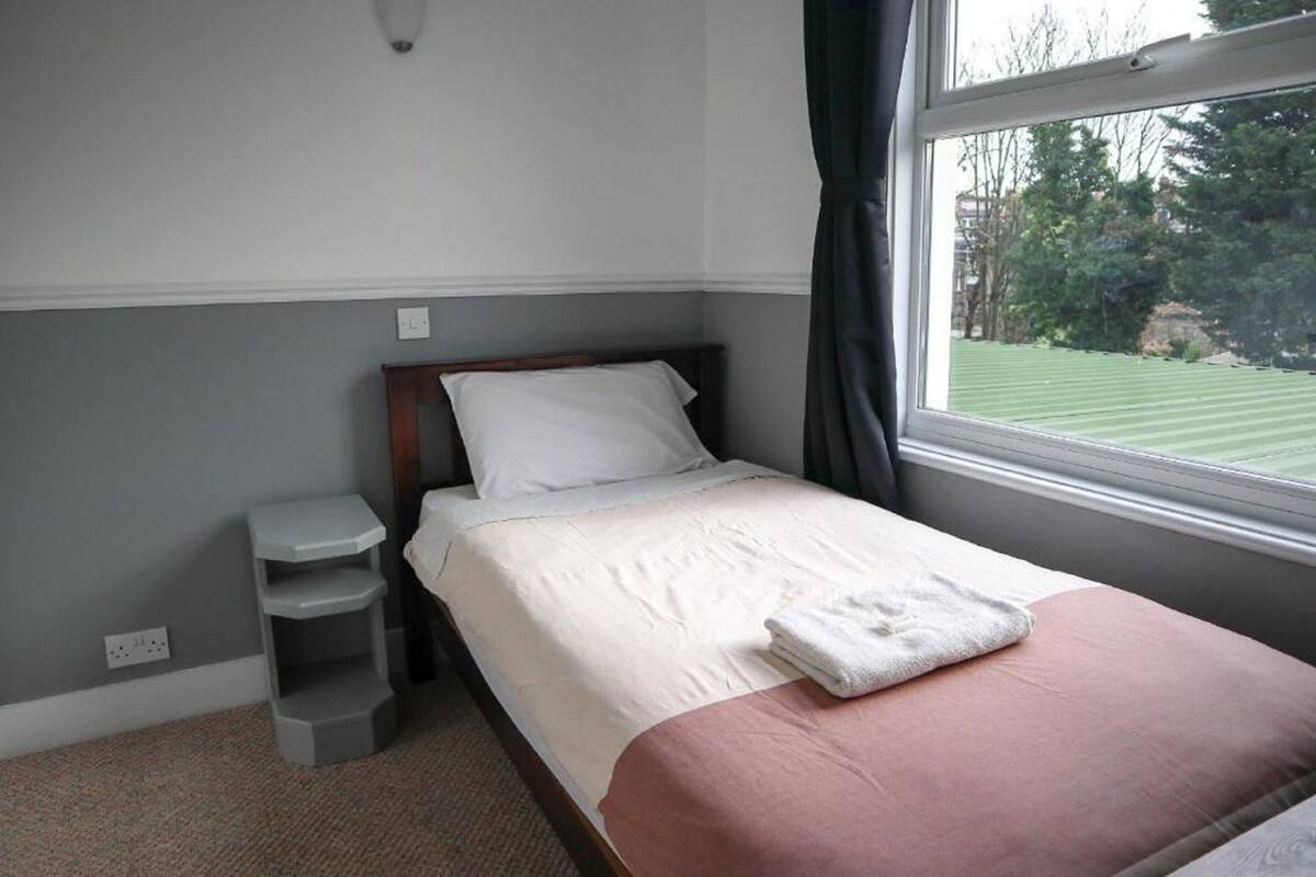A neatly made bed with a white and pink bedspread, a small bedside table, and a window with a view of a green lawn.