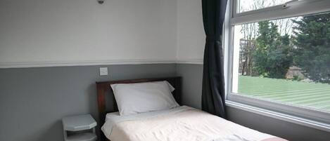 Un lit bien fait, recouvert d’une couvre-lit blanche et rose, une petite table de chevet et une fenêtre donnant sur une pelouse verdoyante.
