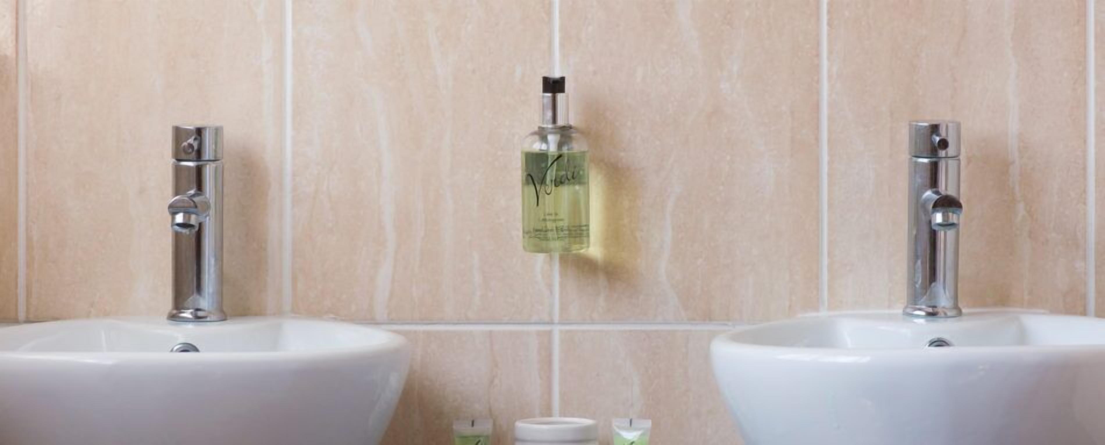 Dos lavamanos blancos con grifos cromados, una botella de jabón para manos y una pequeña botella de loción en la pared.