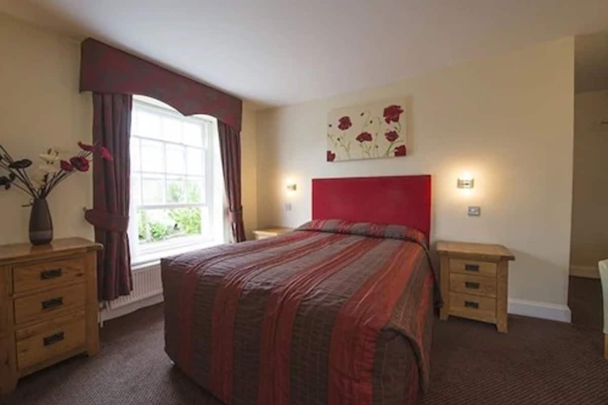 A bedroom with a large bed, a window with curtains, a bedside table, and a wall art piece.