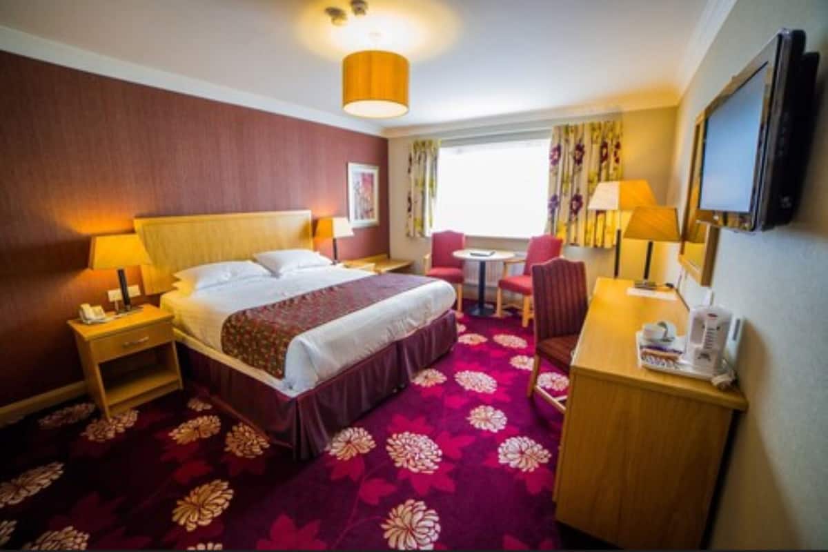 A hotel room with a bed, bedside tables, a desk with a chair, a TV, and a window with curtains.