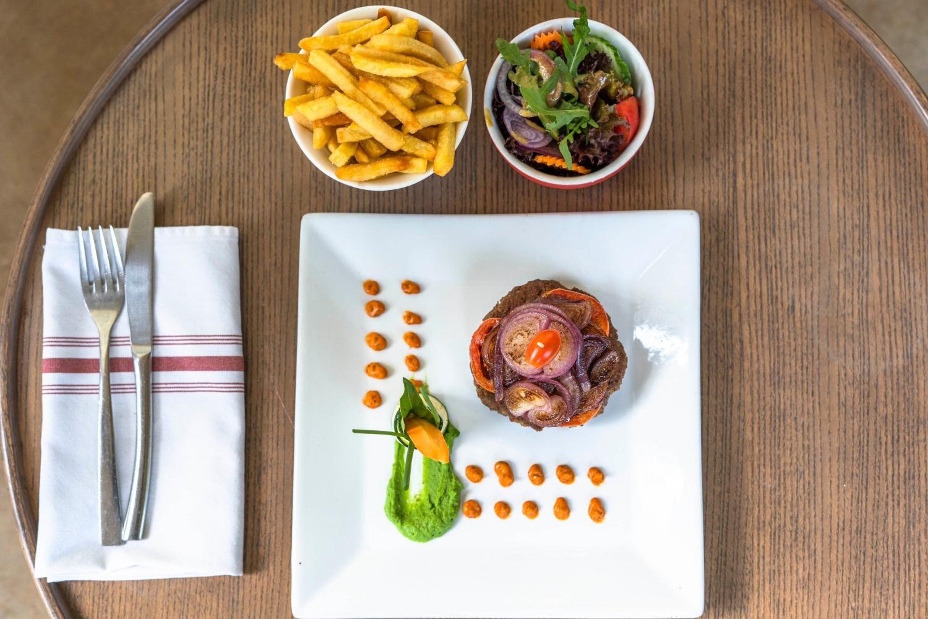 A plate with a steak topped with red onion slices and a garnish, accompanied by a small bowl of fries and a salad on a wooden table.