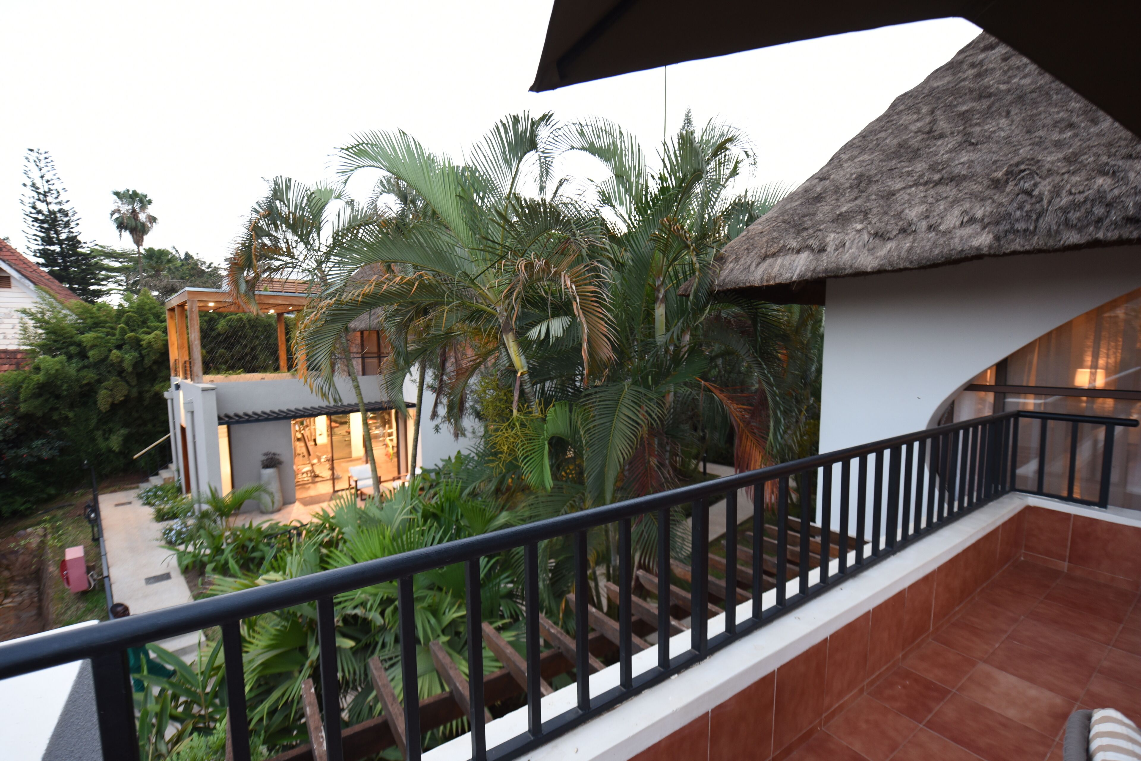 Un balcón con techo de paja, baranda de madera y vista a una casa con patio y palmeras.