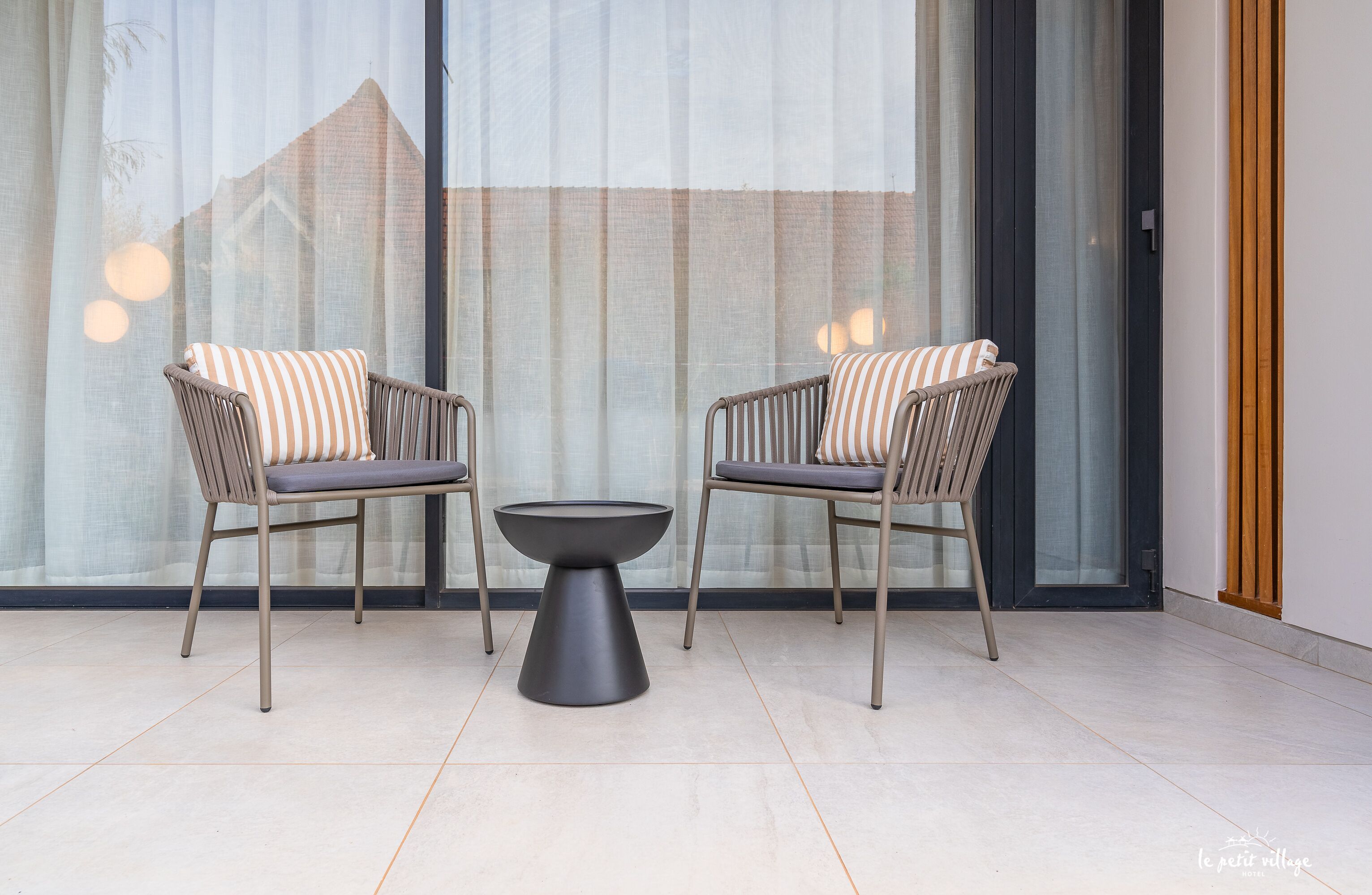 Un espace de détente extérieur moderne, composé de deux fauteuils en osier et d’une table centrale, installé devant une grande fenêtre ornée de rideaux légers.