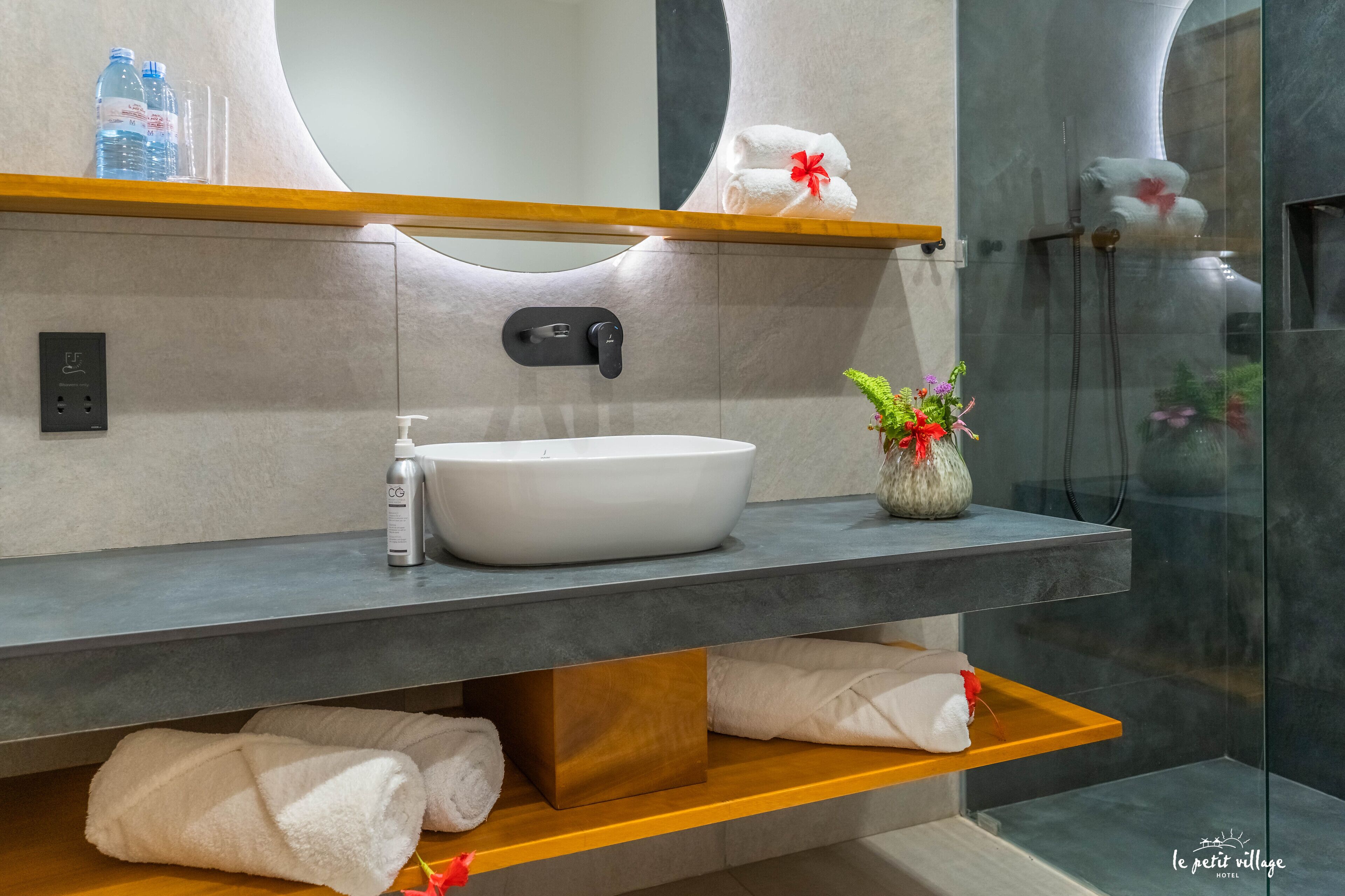 A modern bathroom with a large white sink, a mirror, and a shower area. There are towels and toiletries on the shelves.