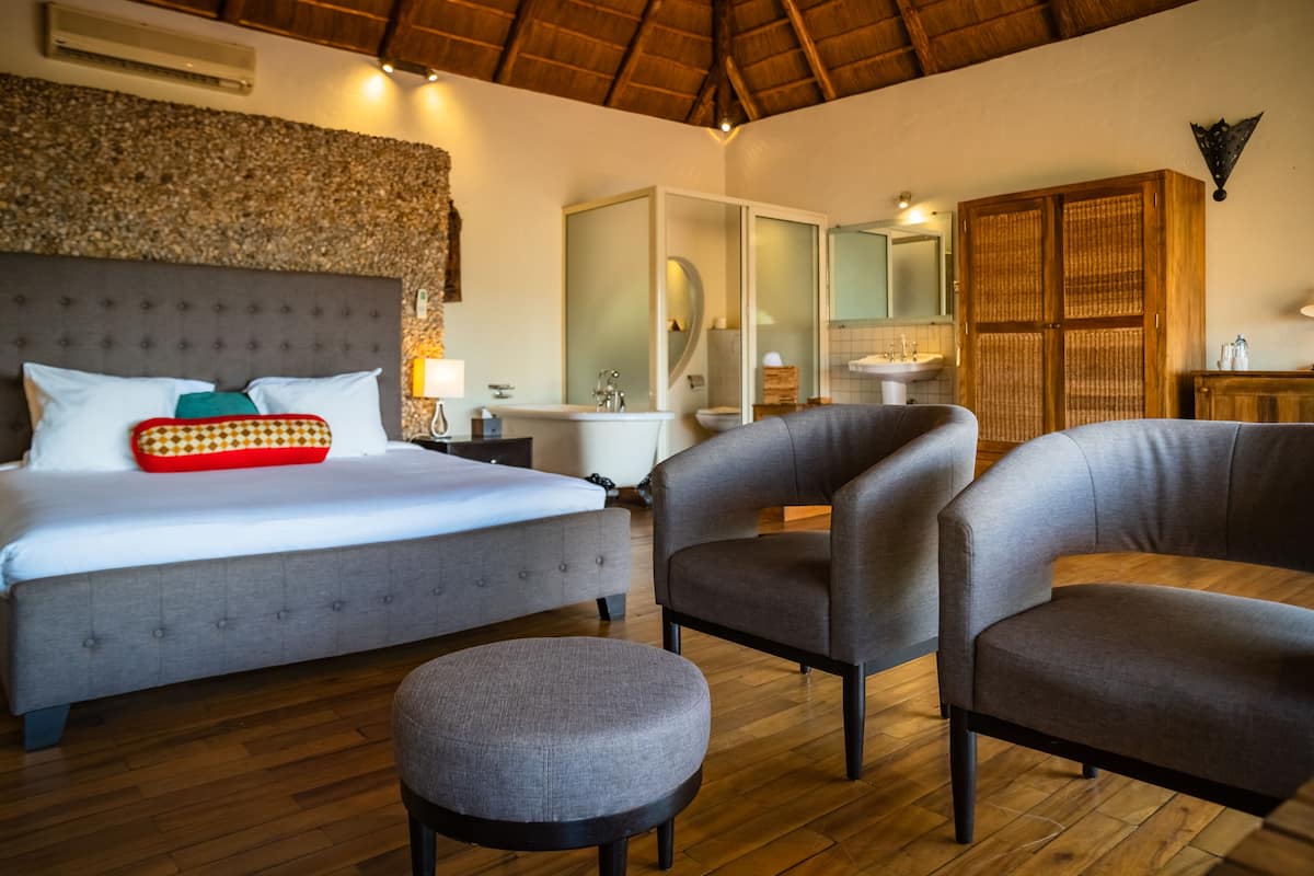 A bedroom with a large bed, two armchairs, a round ottoman, a wooden floor, a bathroom with a bathtub, and a mirror.