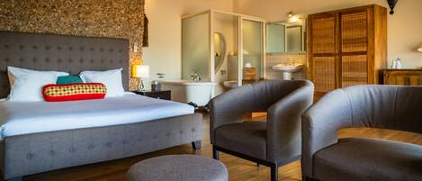 A bedroom with a large bed, two armchairs, a round ottoman, a wooden floor, a bathroom with a bathtub, and a mirror.