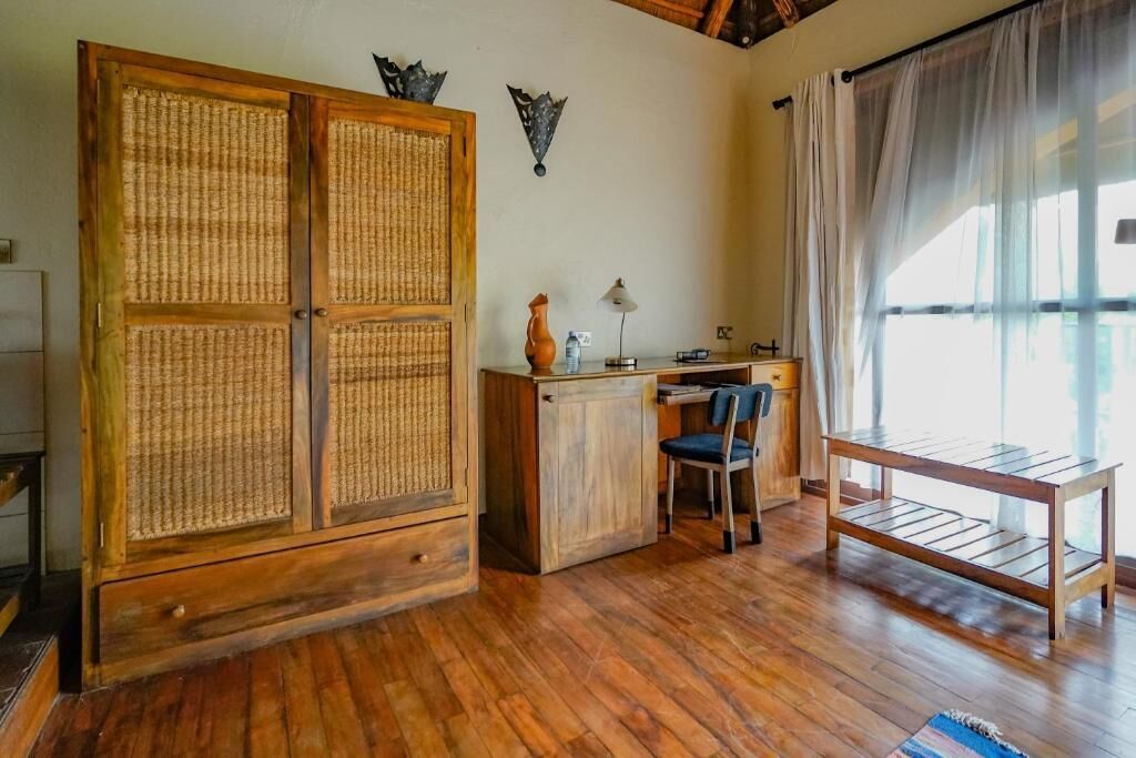 A room with a wooden wardrobe, a desk with a chair, and a window with sheer curtains.