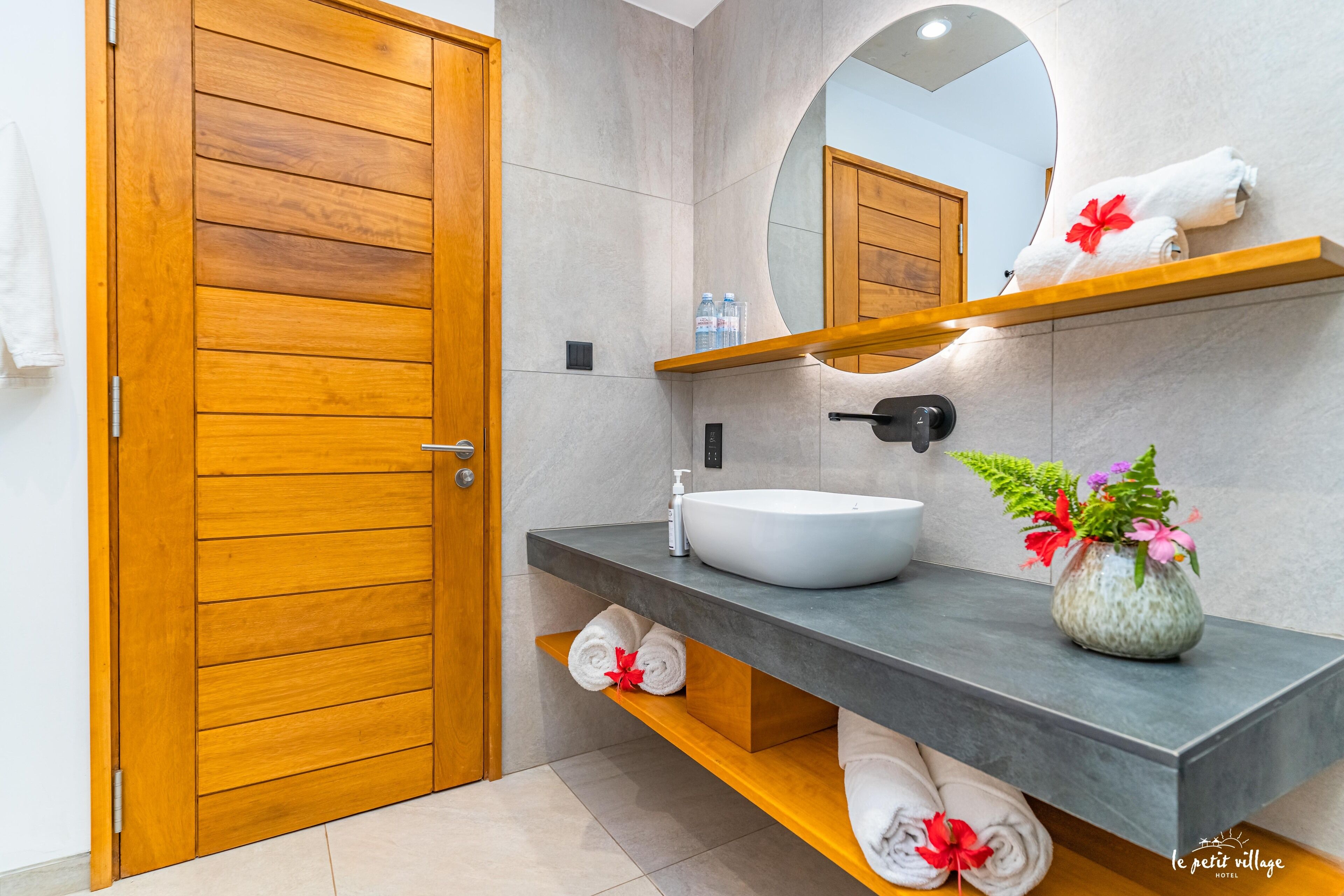 Une salle de bain moderne avec une porte en bois, un miroir rond, un lavabo blanc et une étagère avec des serviettes et un vase de fleurs.