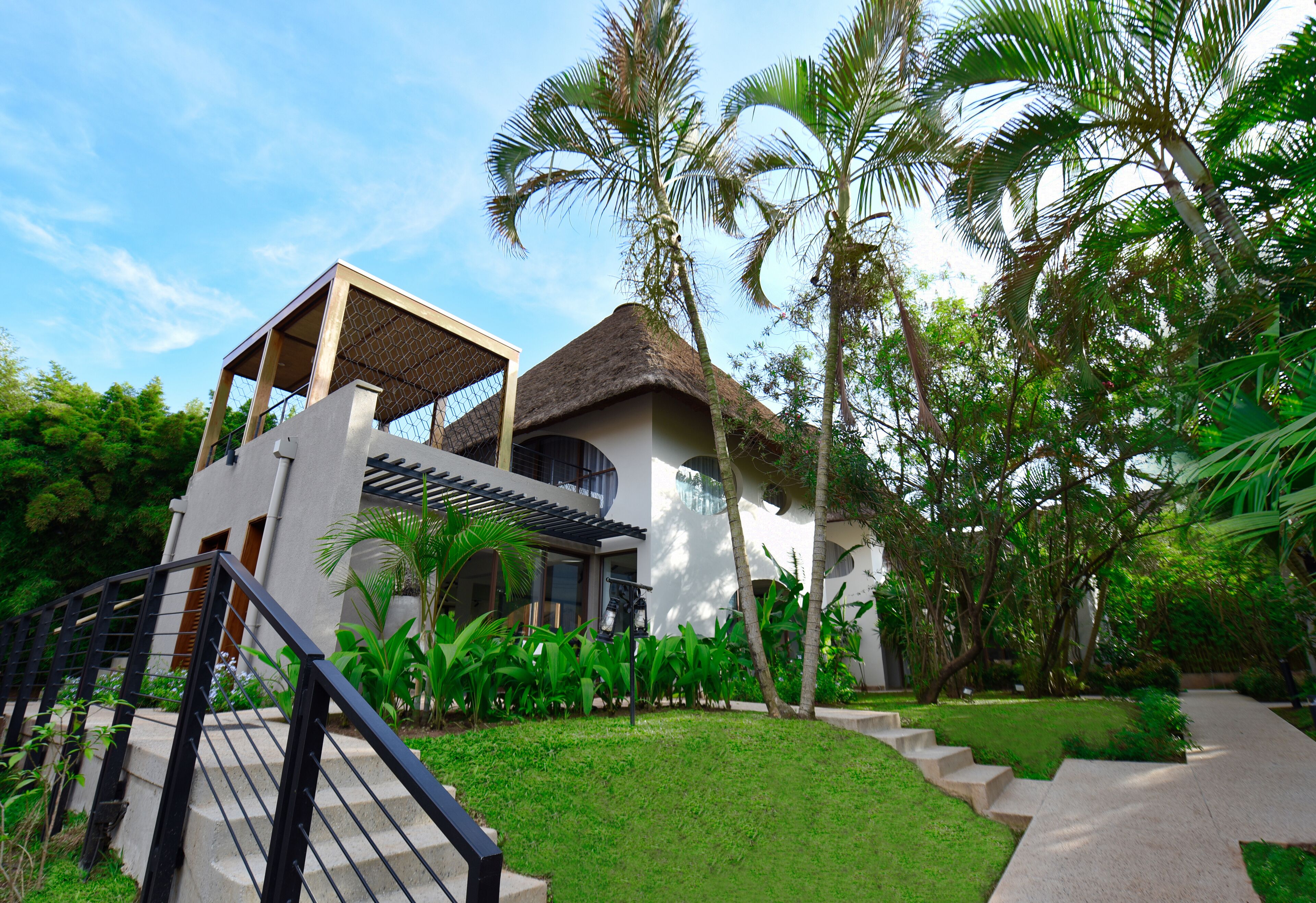 A modern villa with a thatched roof, surrounded by lush greenery and palm trees.