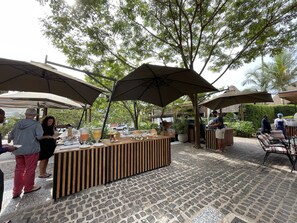 An outdoor dining area with a wooden table and benches, surrounded by umbrellas and greenery. - Le Petit Village Hotel & Spa (Kampala)