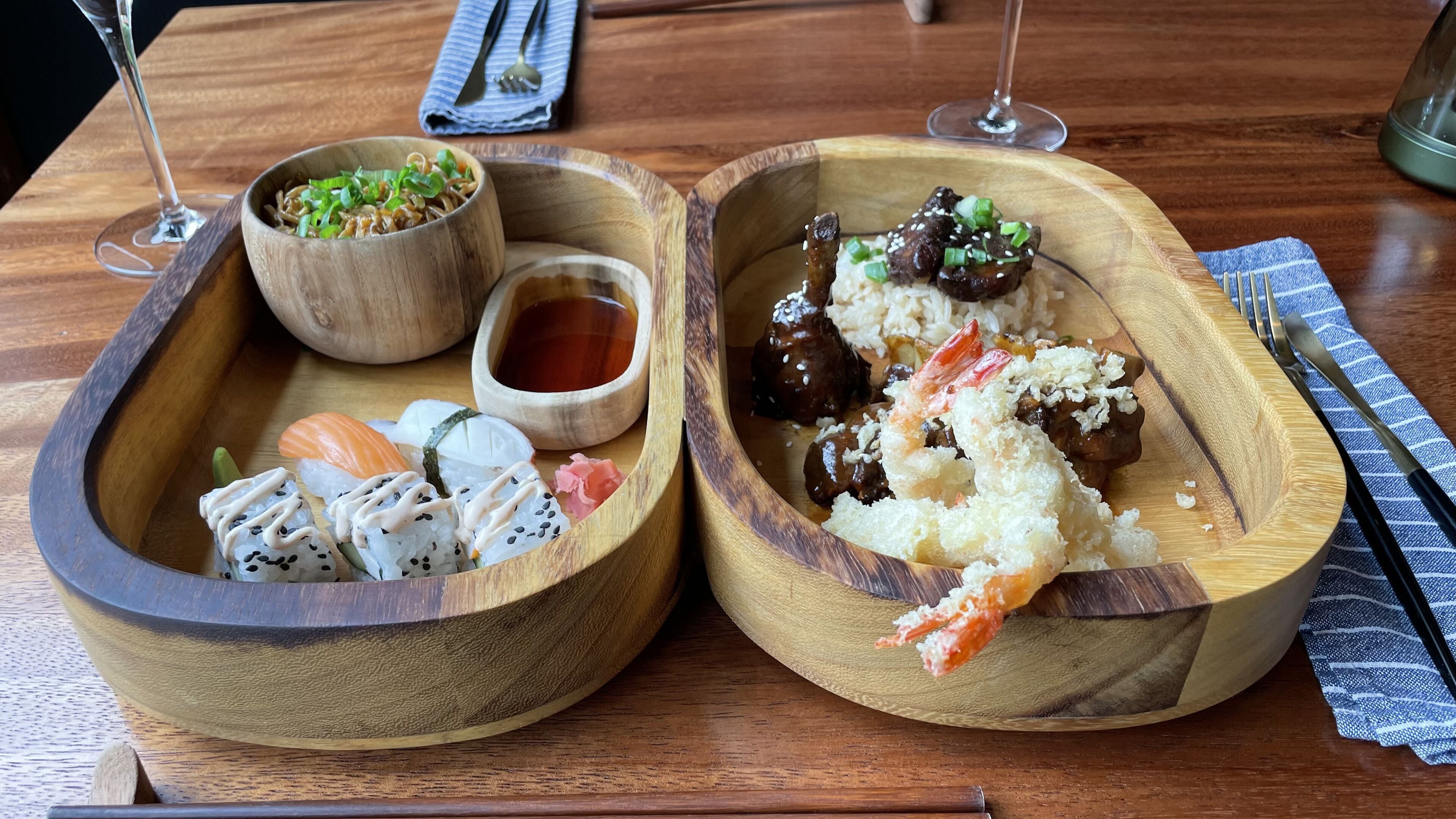 A wooden table with two wooden bowls containing sushi and rice dishes, accompanied by small bowls of sauce and chopsticks.