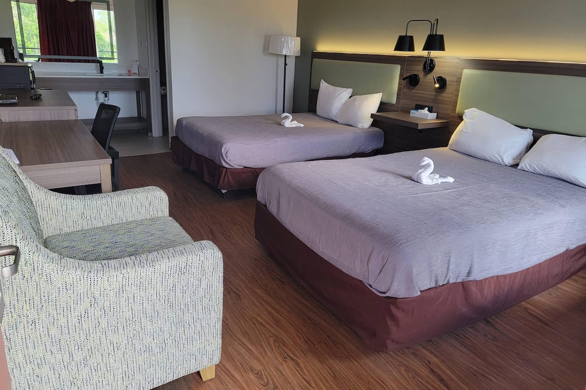 A hotel room with two beds, a desk, a chair, and a lamp.