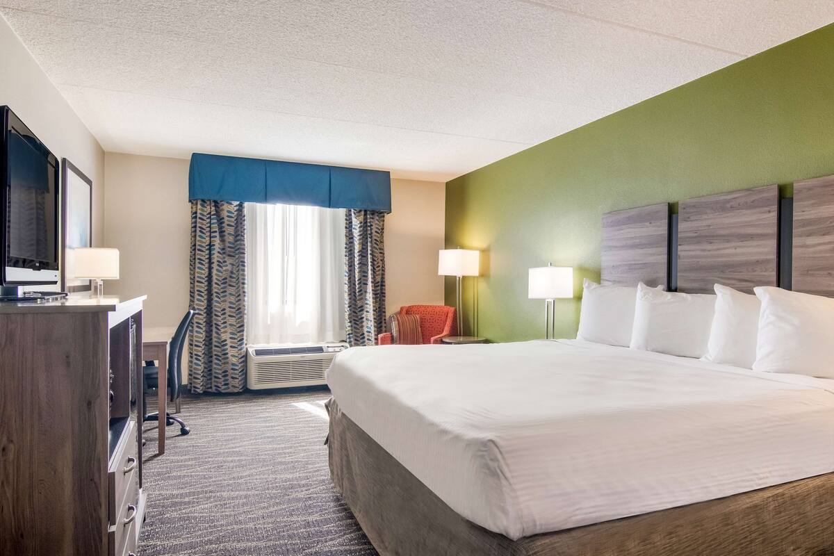 A hotel room with a large bed, two bedside lamps, a desk with a computer, a chair, and a window with curtains.