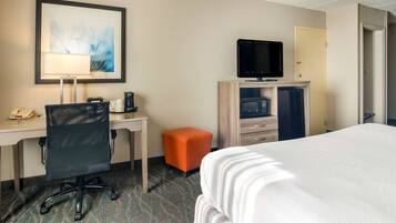 Habitación de hotel con cama, escritorio, silla, televisor, microondas y un pequeño taburete naranja.