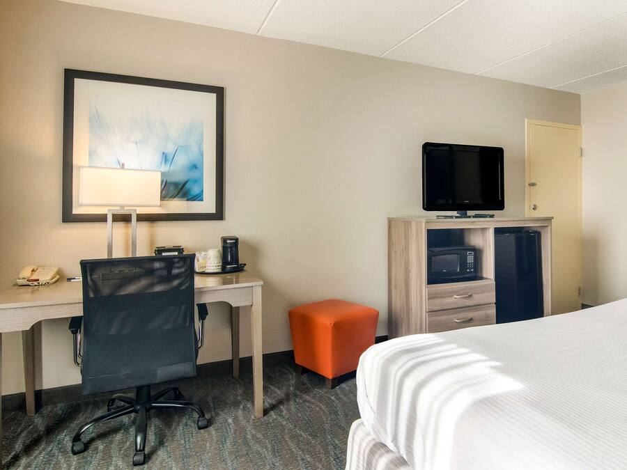 Habitación de hotel con cama, escritorio, silla, televisor, microondas y un pequeño taburete naranja.